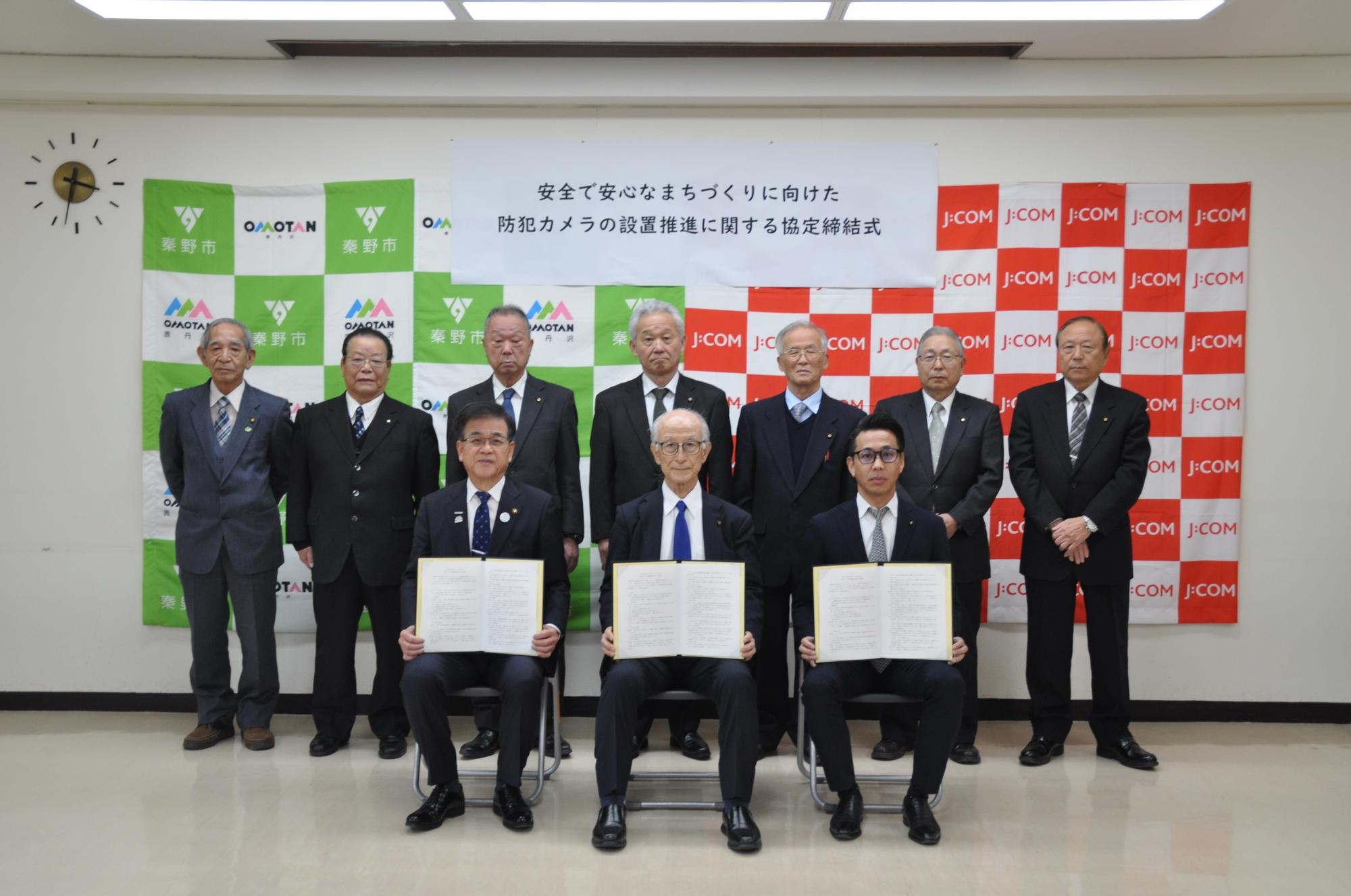「安全で安心なまちづくりに向けた防犯カメラの設置推進に関する協定締結式」と書かれたボードを背景に、3名が協定書を手に座り、その後ろに7名のスーツ姿の男性が並んで立っている記念写真