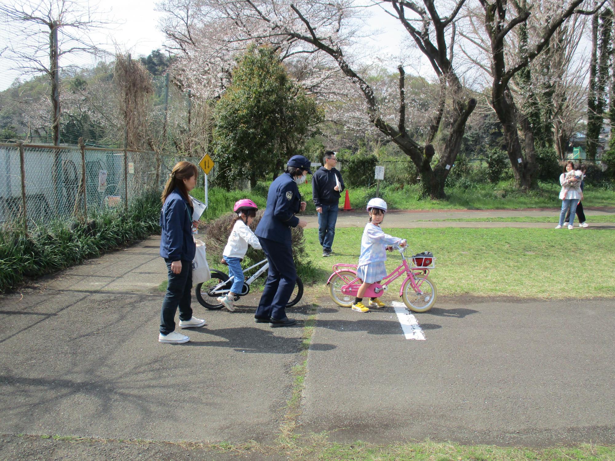 桜が咲く公園の舗装された通路で、制服を着た警察官がヘルメットを被った小さな子供2人を含む数人のグループに対し、ピンク色の自転車に乗った子供に乗り方を指導している交通安全教室の様子の写真