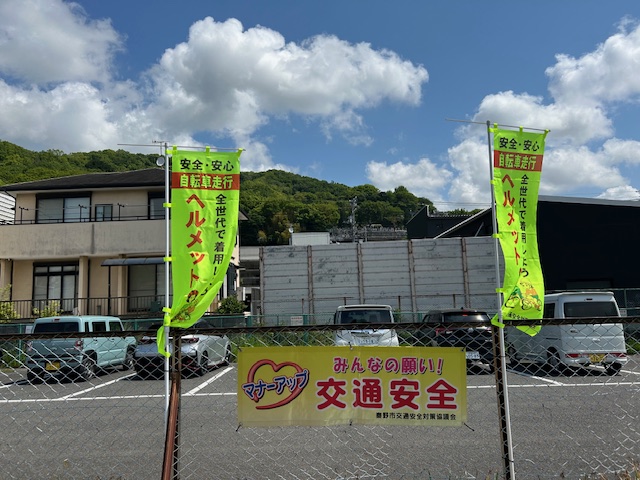 駐車場のフェンスに「みんなの願い！交通安全」の横断幕が掲げられ、「安全・安心自転車走行 全世代で着用しよう ヘルメット着用！」と書かれた蛍光グリーンののぼりが2本立っている写真
