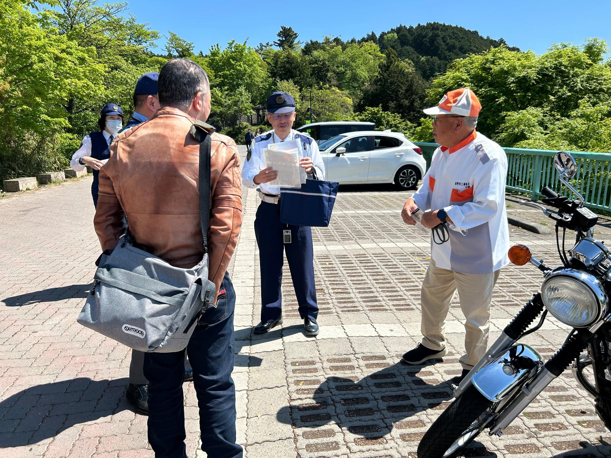 警察官と交通安全運動の帽子をかぶった男性を中心に、数人が集まり、書類のようなものを確認しながら立ち話をしている様子の写真