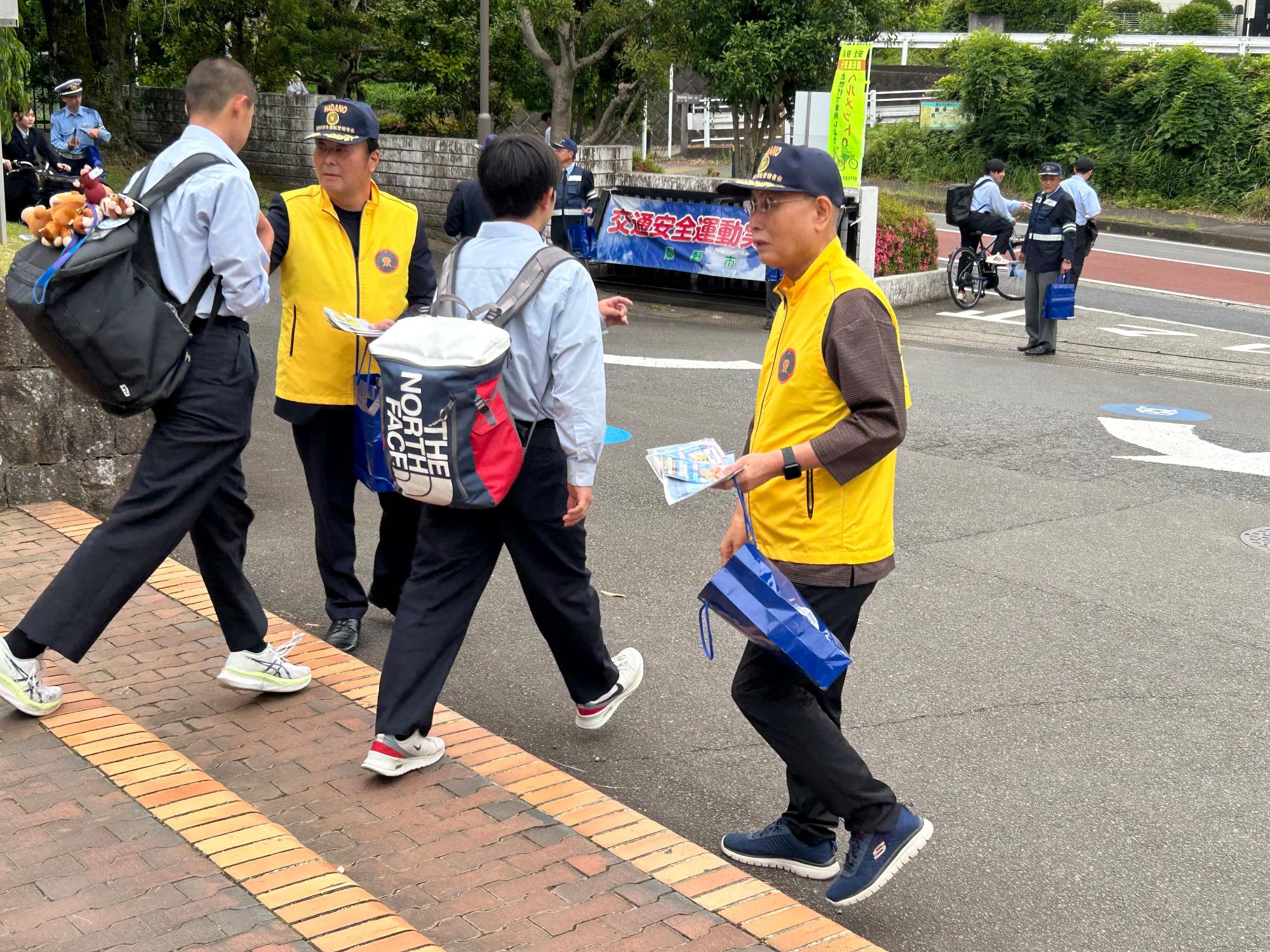 制服を着た警察官と黄色のベストを着用した市民らが、登校中の学生に青い手提げ袋とチラシのようなものを渡しながら交通安全を呼びかけている様子の写真