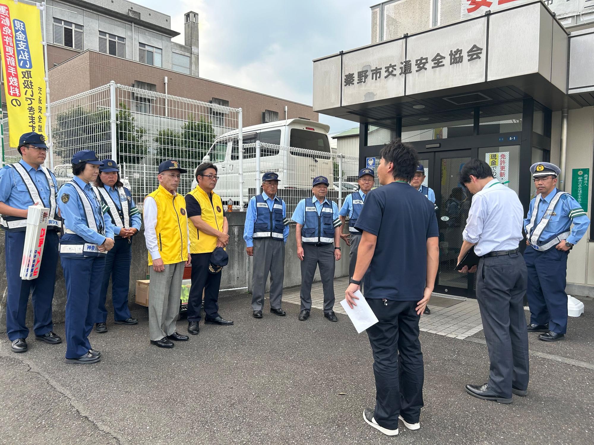 曇りの日に、「秦野交通安全協会」と書かれた建物の前で、制服の警察官や黄色と青色のベストを着た市民らが集まり、手前の男性の指示を聞いている様子の写真