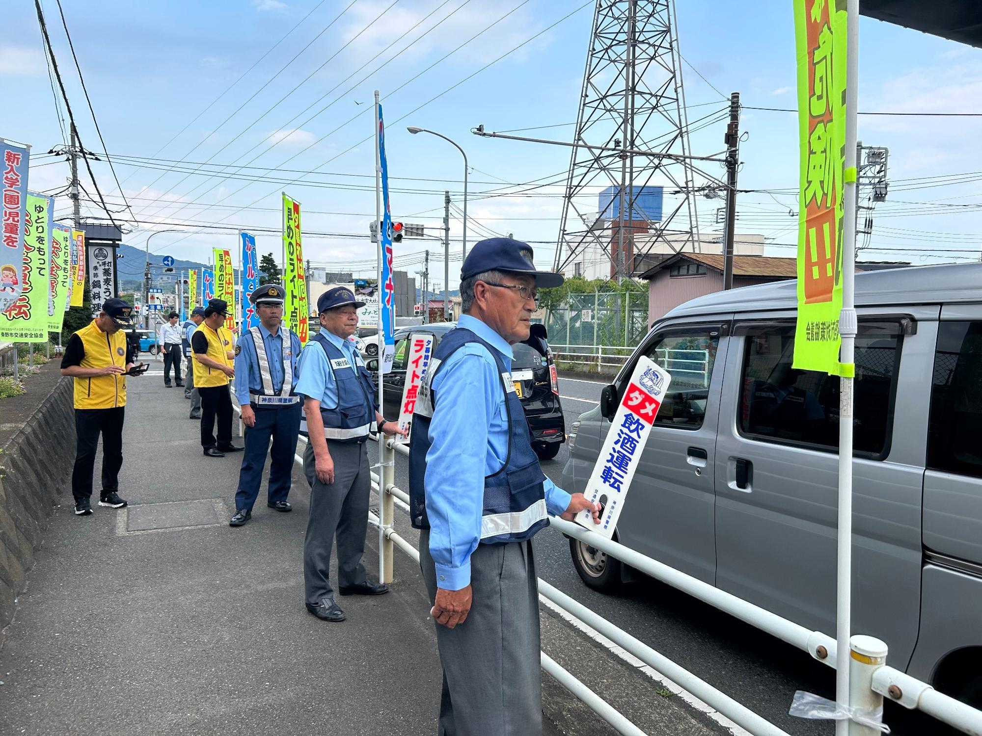 車道脇の歩道で、制服の警察官や黄色のベストを着用した市民らが、飲酒運転根絶ののぼりやプラカードを持ち、一列に並んでドライバーに交通安全の啓発活動をしている様子の写真