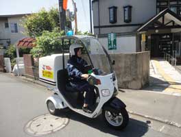 三輪の屋根付き青パトバイクに乗った制服姿の配達員が、住宅地の舗装された道路を走行している写真