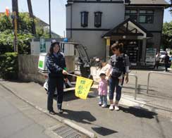 駐車した青パトバイクのすぐ横で、制服を着た人が黄色い旗を持ち、見守り活動を行っている様子の写真