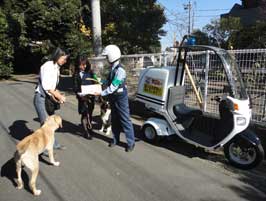 配達員が屋根付きの青パトバイクの横に立ち、大型犬を散歩中の女性たちと話をしている様子の写真