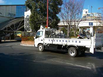 荷台に放置自転車が大量に積まれ、赤色灯を装備した白と黒の小型トラックが回収する様子の写真