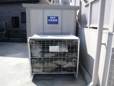 白い防水シートで覆われたメッシュ構造の金属製防災倉庫が屋外に設置され、青い「備蓄用発電機置場」の案内板が掲示された災害対策施設の写真