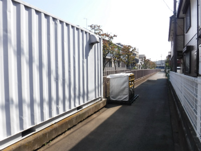 住宅街の細い道路脇に設置された銀色の縦縞模様を持つ大型防災用コンテナ倉庫が、塀と接して配置されいる写真