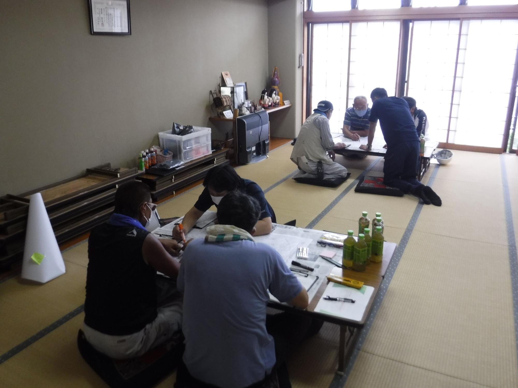 和室の畳の上で、マスクを着用した数名の男性が二つの座卓に分かれ、それぞれ地図のような紙や資料を広げて、防災対策に関する意見交換や作業を行っている写真