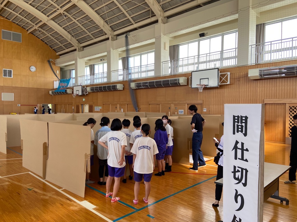 体育館で生徒たちが段ボールの簡易間仕切りを組み立てる訓練を行っており、手前には「間仕切り」と書かれた大きな案内板がある、防災訓練の様子の写真