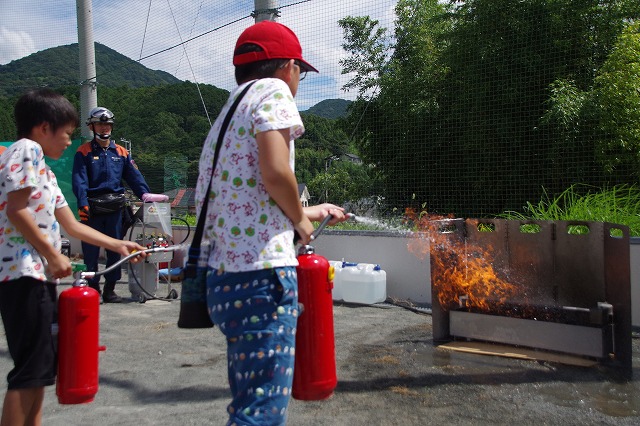野外の広場で、二人の子どもが赤い消火器を操作して、炎が上がっている訓練用の火元に放水し、後ろでは消防隊員の男性が見守っている消火訓練の写真