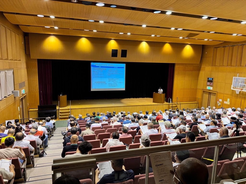 木目調の天井と暖色照明が特徴の大規模ホールで、多数の参加者がエンジ色の座席に着席しステージ上の青いスクリーン投影資料を見る研修会の写真