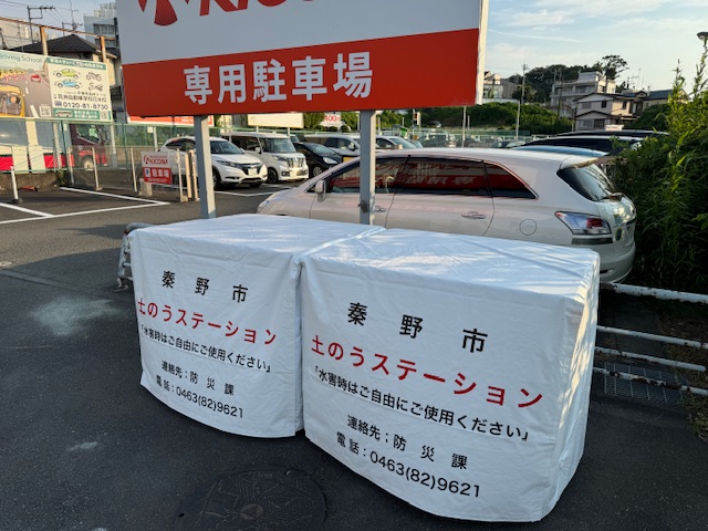 スーパーの専用駐車場前に「秦野市 土のうステーション」と印字された二基の白いコンテナが設置され、市民が自由に防災用土嚢を利用できる常設配布拠点の写真