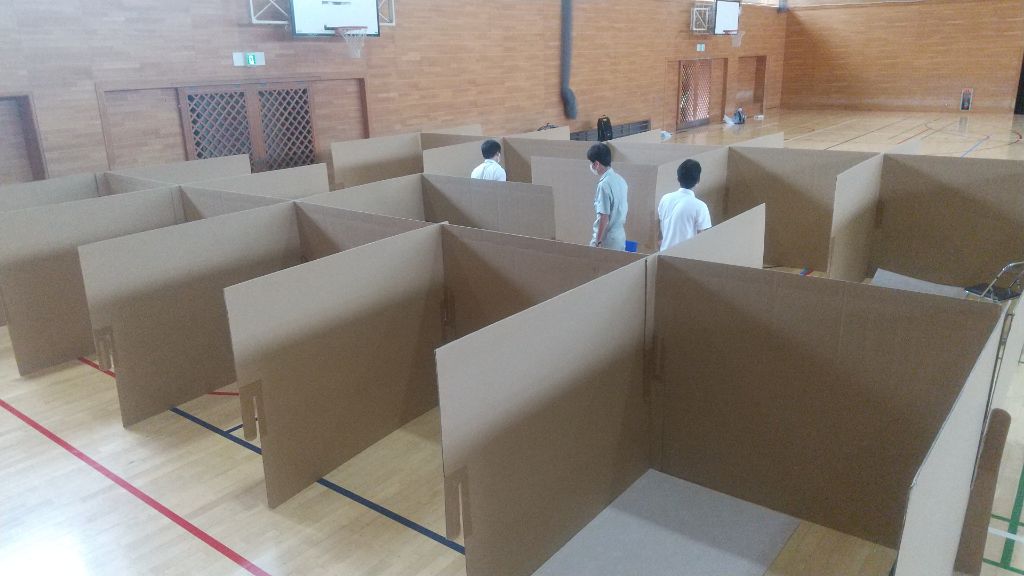 育館の床に段ボールで組み立てられたパーティションが設置され、プライバシー保護のために仕切られた空間の中に数名の人が立っている写真
