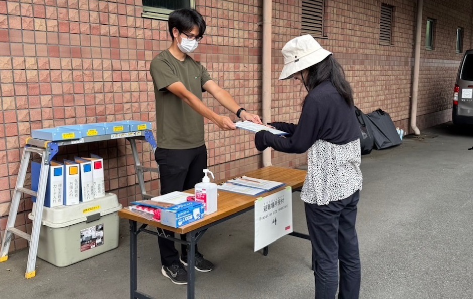 屋外の受付らしき場所で、マスクを着けた男性スタッフが、帽子をかぶった女性に書類を手渡しており、テーブルの上には消毒液と「避難者受付」と書かれた貼り紙がある災害対応の写真
