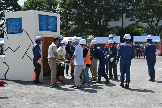 ひび割れが描かれた建物模型の前で、白いヘルメットを着用した参加者が協力し、消防隊員の指導の下、担架を使って負傷者を救助する訓練を行っている防災訓練の写真
