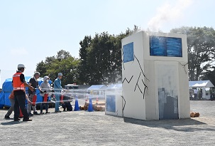 数名の人々が放水訓練に参加し、ひび割れが描かれた建物模型の窓から噴き出す煙めがけて消防ホースで勢いよく水を放っている訓練風景の写真