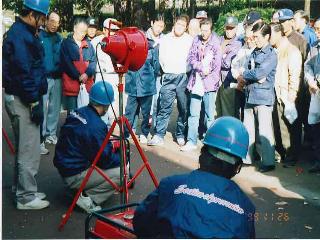公園などの屋外で消防署員がヘルメットを着用し消火器を使った初期消火訓練を実演し、多数の地域住民が円形に囲んで見学する防災啓発活動の写真