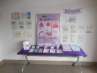 白い壁に「勇気はYESと思い込むのはNO! 相手の言動が怖いと思ったら連絡力を」と紫色で書かれた大型DV防止ポスターと相談窓口案内を掲示し、紫布のテーブルに多数のパンフレットを配架した女性支援啓発コーナーの写真