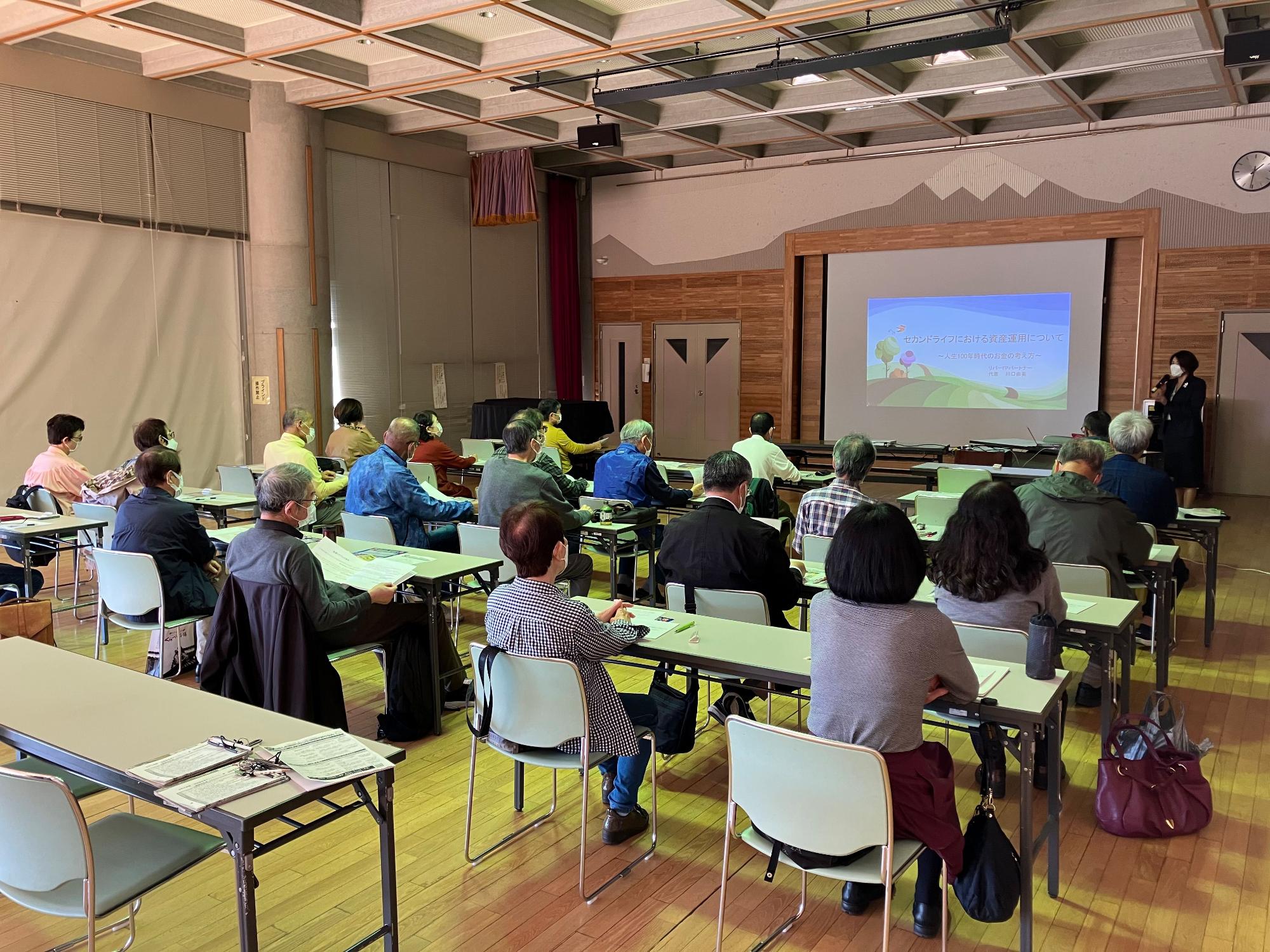 木目調の壁と山並みのシルエットが特徴的な多目的ホールで、黄緑のテーブルに着席した参加者が移住支援担当者のプレゼンテーションを聞いている様子を後方から写した写真