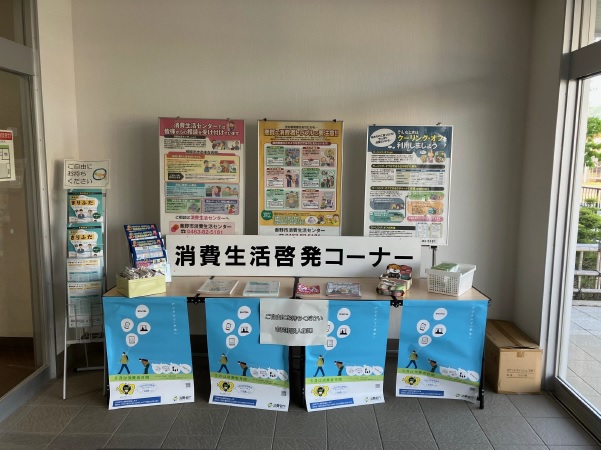 明るい窓際に設置された「消費生活啓発コーナー」に数種類の啓発ポスターが並び、4枚のポスターが貼られた台には複数のパンフレットやチラシが並べられている写真