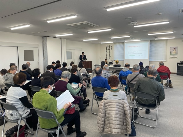 白い壁と蛍光灯照明の会議室で、スーツ姿の講師が演台とスクリーンを使いプレゼンテーションを行い、多数の参加者が椅子に座って資料を手に聞き入る講演会の写真