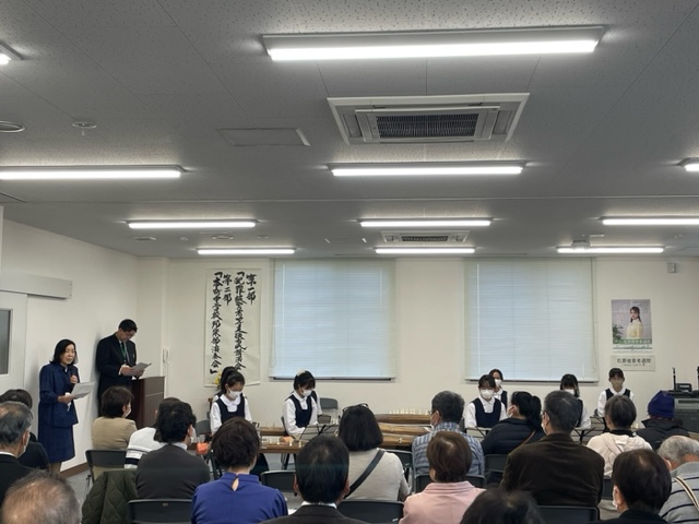 会議室で行われている発表会の様子で、演台に立つ二人の司会者と中央に並ぶ白いベストを着た複数の発表者、そして多くの聴衆が着席している室内の写真