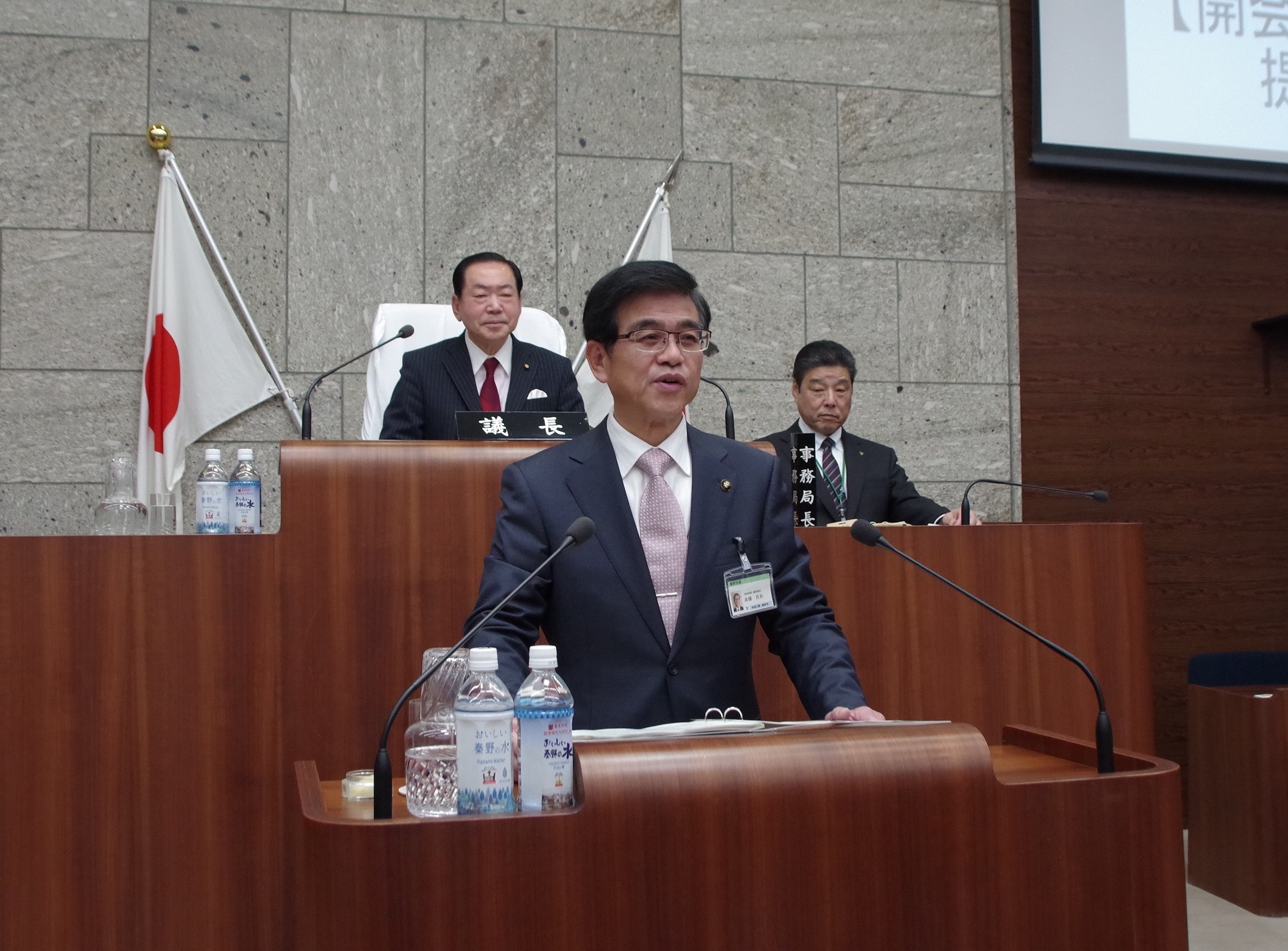 和装のような石造りの壁の議場内で、ダークスーツに薄いピンクのネクタイを締めた市長が木製の演壇で発言しており、その後ろには日章旗と、それぞれ「議長」「事務局長」と書かれた札の前に着席する2人の男性がいる写真