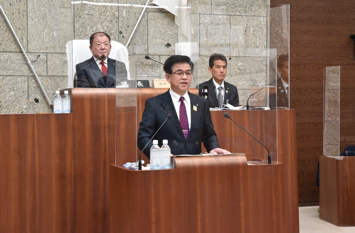 ダークスーツにえんじ色のネクタイを締めた市長が、議会のような場所の木製演壇で透明な仕切り越しに演説しており、その後ろには別の2人のスーツ姿の男性が着席している写真