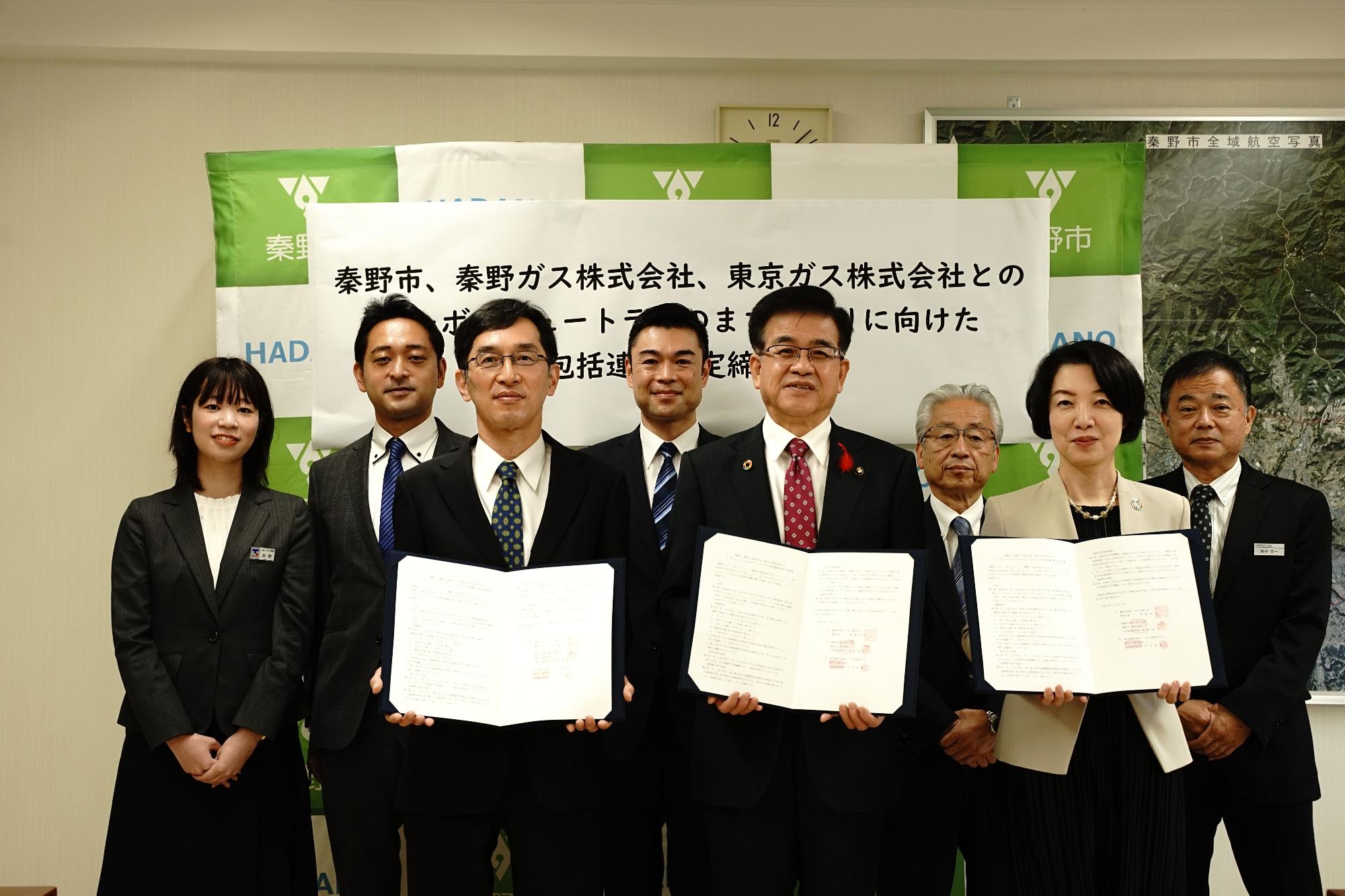 秦野市のバックボードの前で高橋市長と秦野ガス株式会社・東京ガス株式会社の関係者3名が協定書を持ち、その後ろに5名が並び記念撮影をしている写真