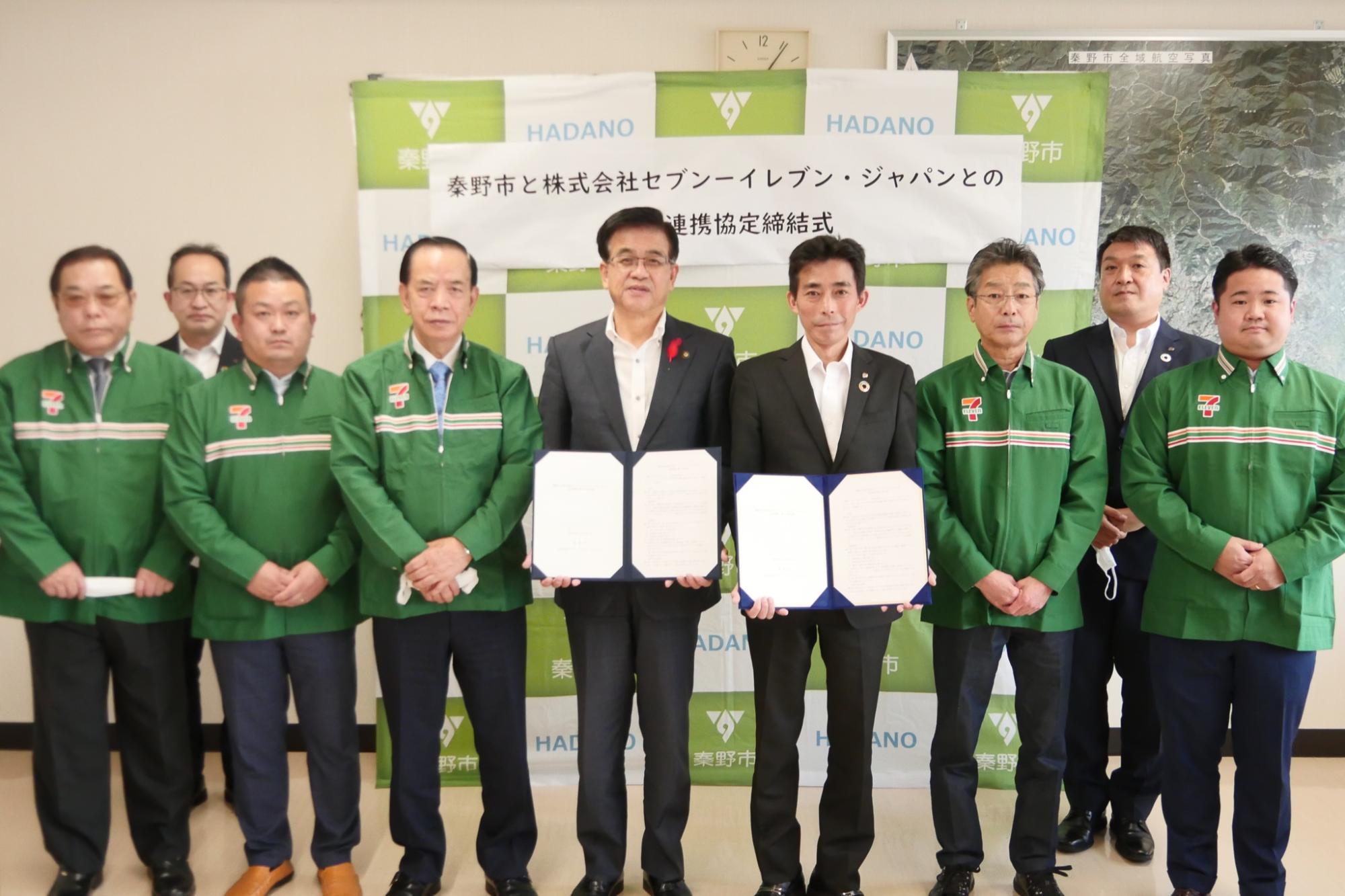 秦野市のバックボードの前で協定書を持った高橋市長と株式会社セブン‐イレブン・ジャパンの関係者が中心に横一列に7名が並び、その後ろに2名の方が並び記念撮影をしている写真
