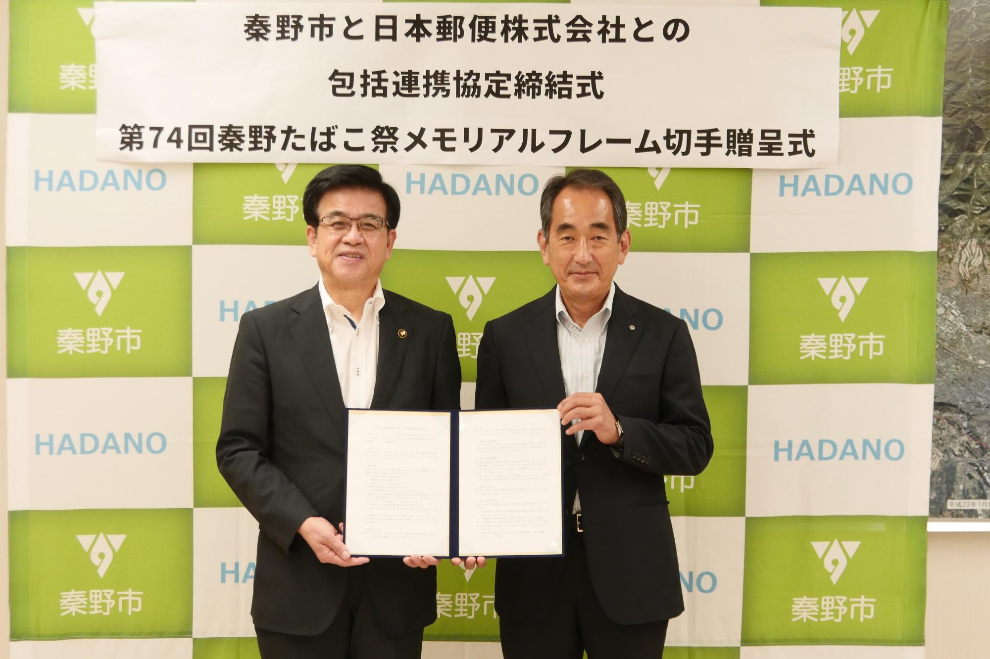 秦野市のバックボードの前で高橋市長と日本郵便株式会社の男性が協定書を持ち記念撮影をしている写真
