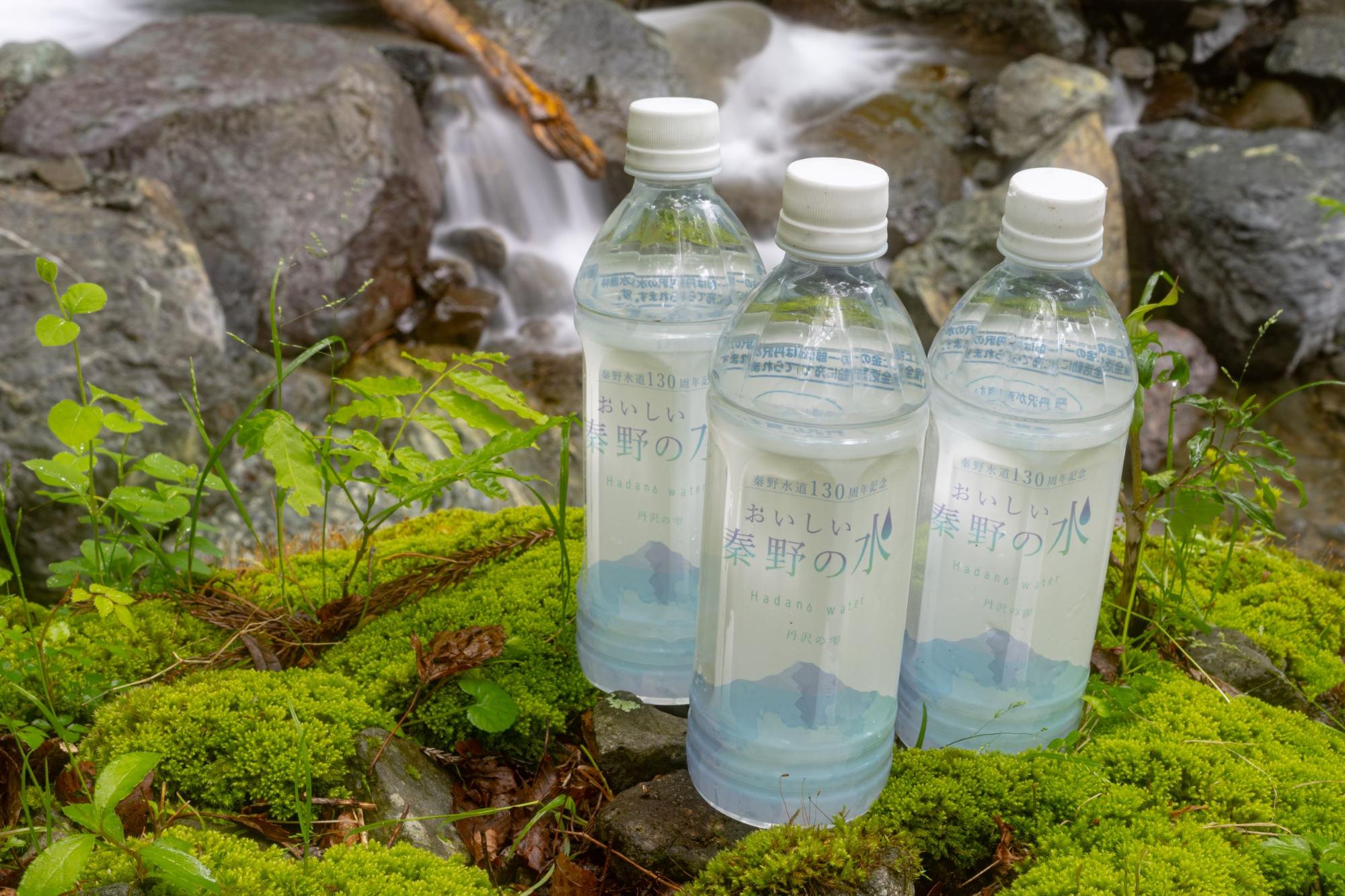 豊かな緑のコケが生い茂る岩場に、水の流れが背景に見える自然の中で、秦野市の水道水を使用した「おいしい秦野の水」と書かれたペットボトル飲料が三本並べて置かれている写真
