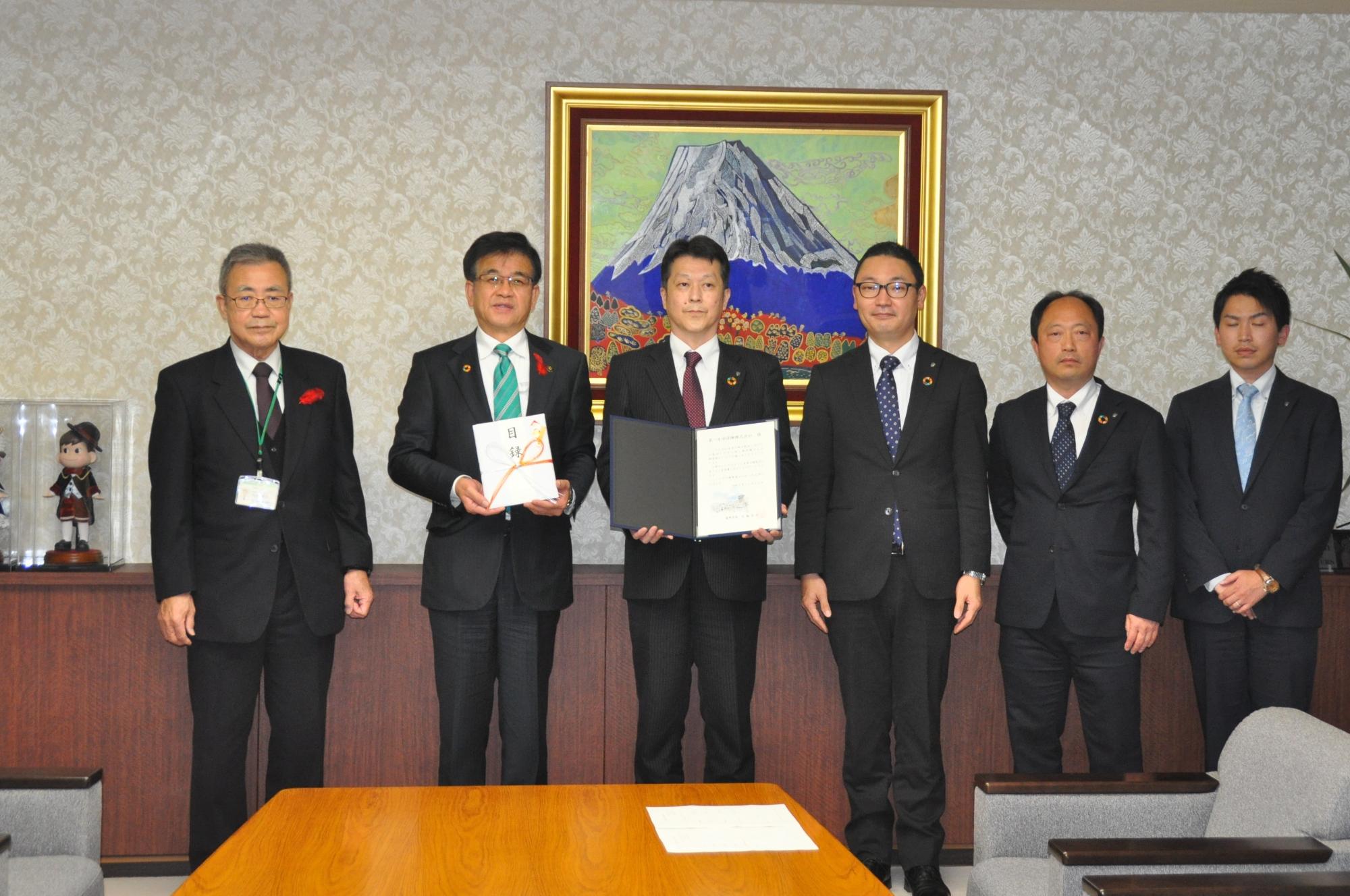 富士山の絵画を背景にした室内のカウンター前で、市長と第一生命保険株式会社の関係者5名で、中央で証書のような書類を広げて持ち、カメラに向かって真剣な表情で立っている記念写真