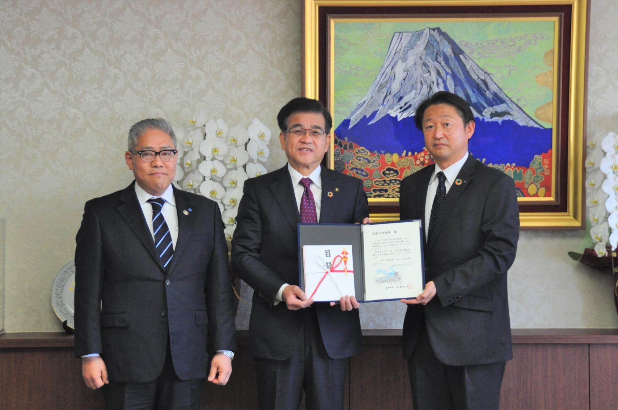 富士山の絵画を背景にした室内のカウンター前で、市長と信金中央金庫の関係者2名が、中央で証書のような書類を広げて持ち、カメラに向かって真剣な表情で立っている記念写真