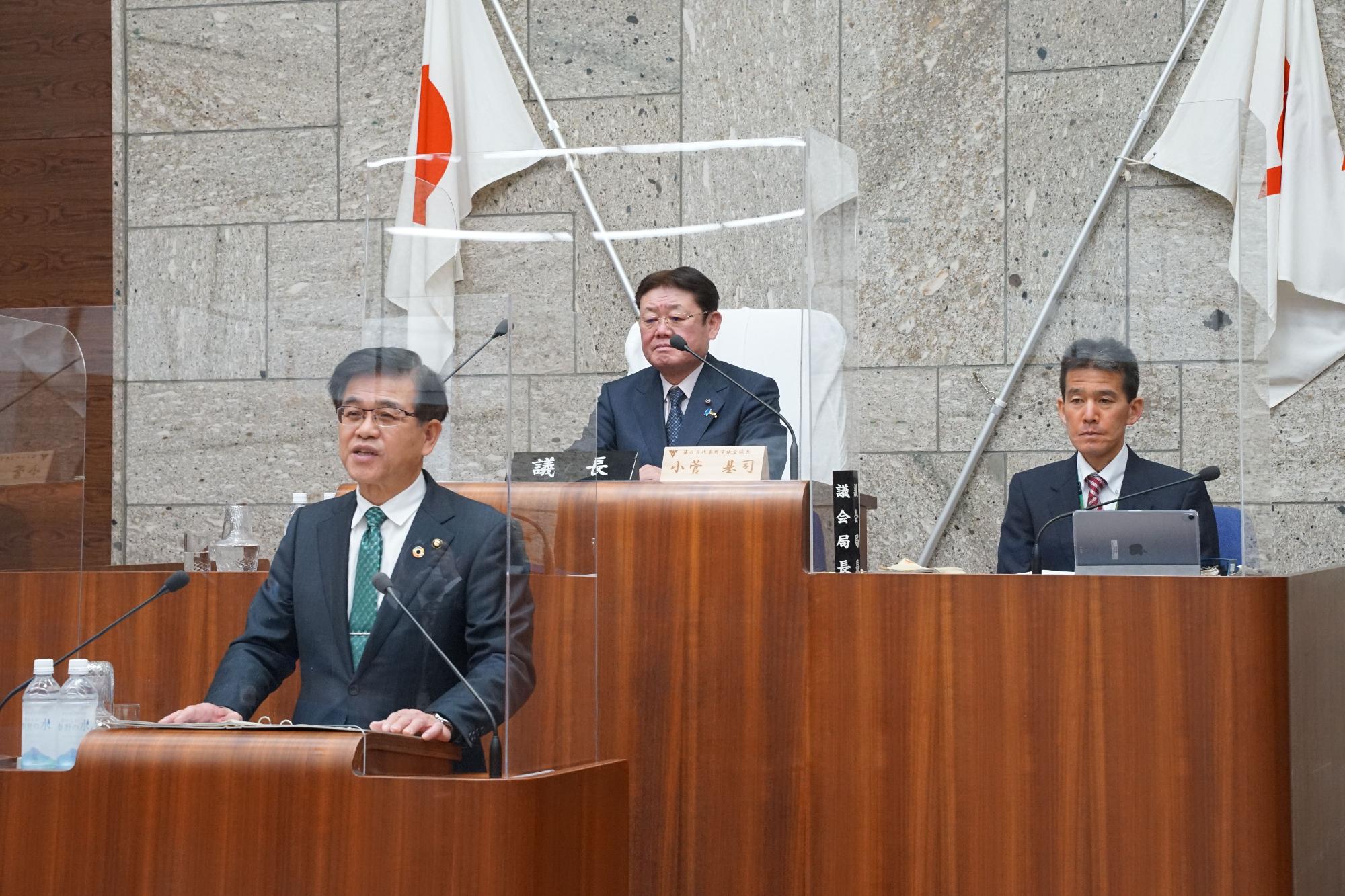 議場でスーツ姿の市長が演壇に立ち発言しており、背後には議長席と日本国旗が二本掲げられ、透明なアクリル板越しに三名の男性が着席している様子を写した公式会議の写真