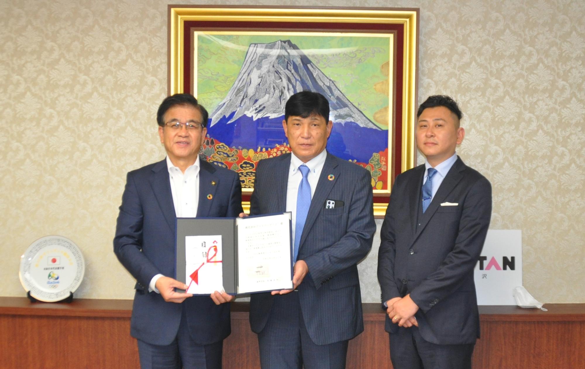 富士山の絵画を背景にしたカウンター前で、市長とスーツ姿の2人の男性が並び、中央と左の男性は証書のような書類と紅白の水引がかかった封筒を持って立っている記念写真