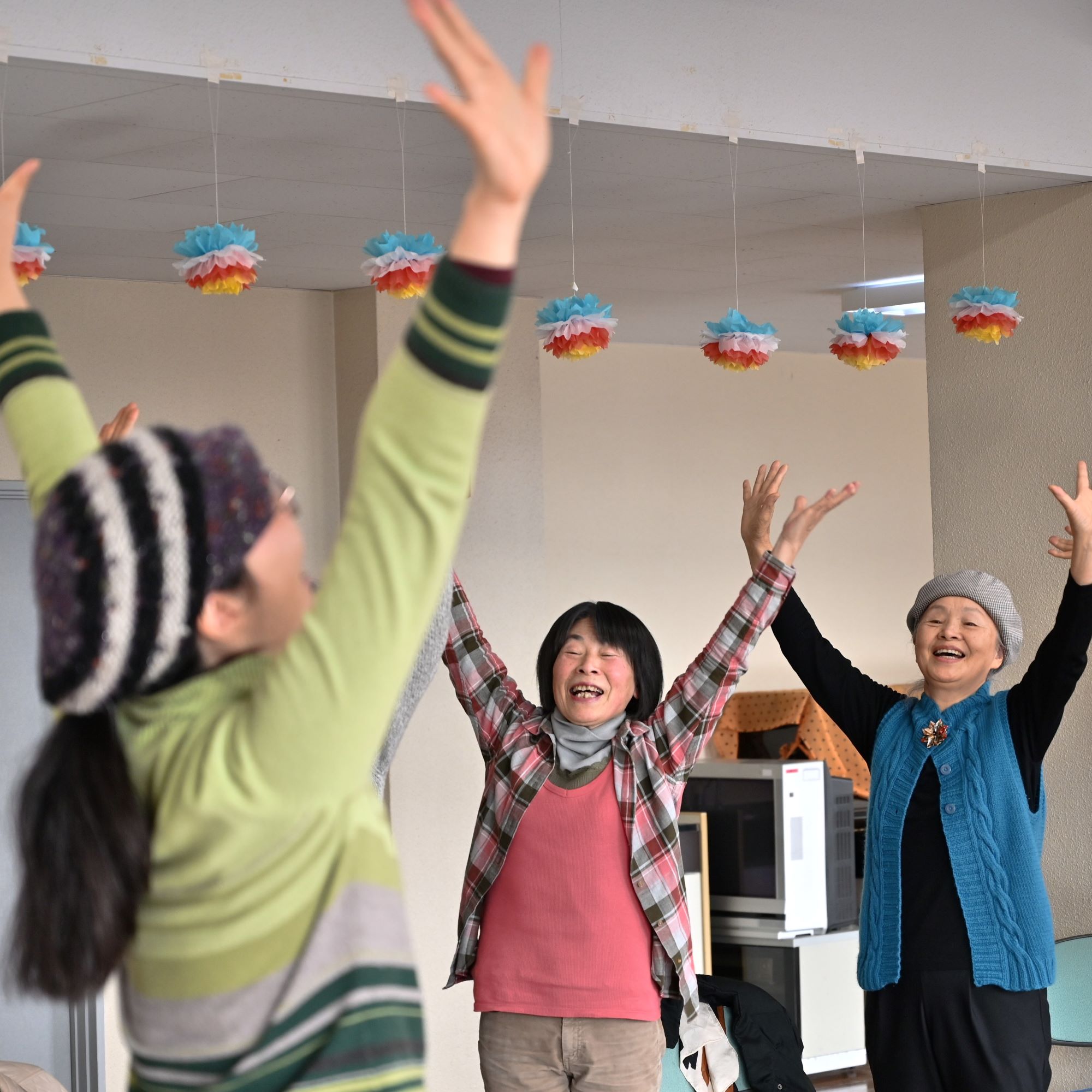 カラフルなボンボン飾りの下で、3人の女性が両手を真上に伸ばして笑顔で楽しく運動やレクリエーションをしている様子を捉えた写真