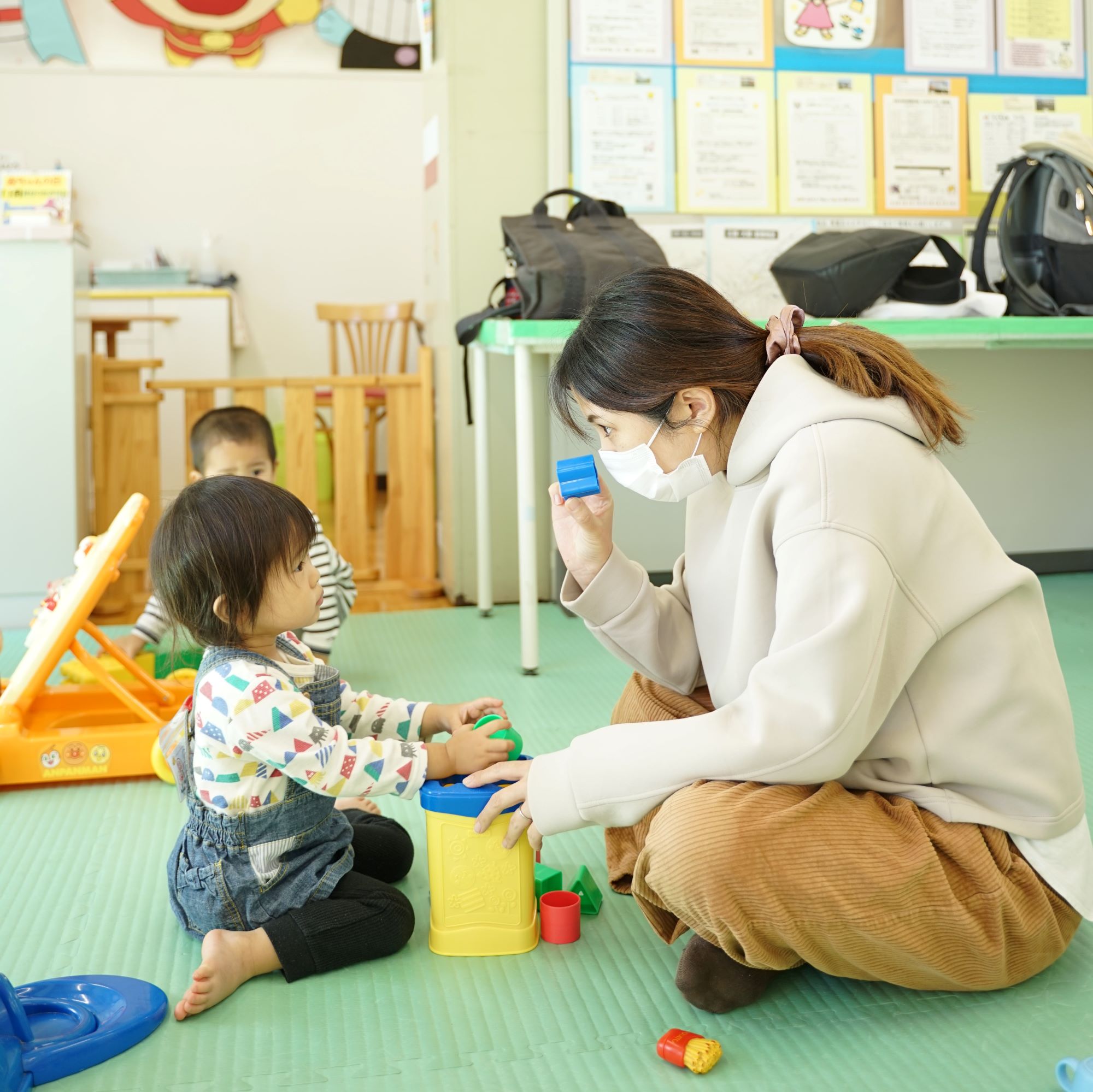 マスクを着用した女性がしゃがみ込み、幼い女の子とブロック遊びを通してコミュニケーションをとっている保育室の写真