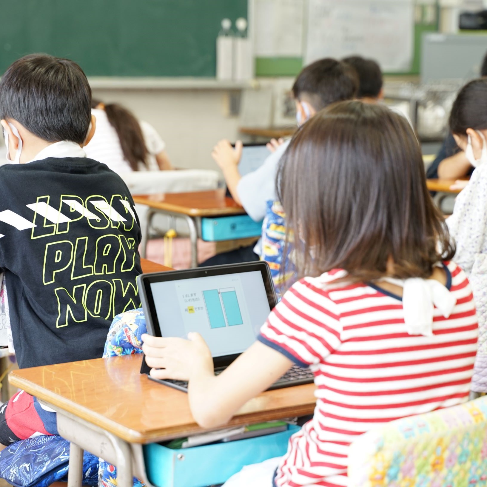 多くの小学生がマスクを着用し、各自の学習机でタブレット型パソコンを使って授業を受けている様子を後方から撮影した写真