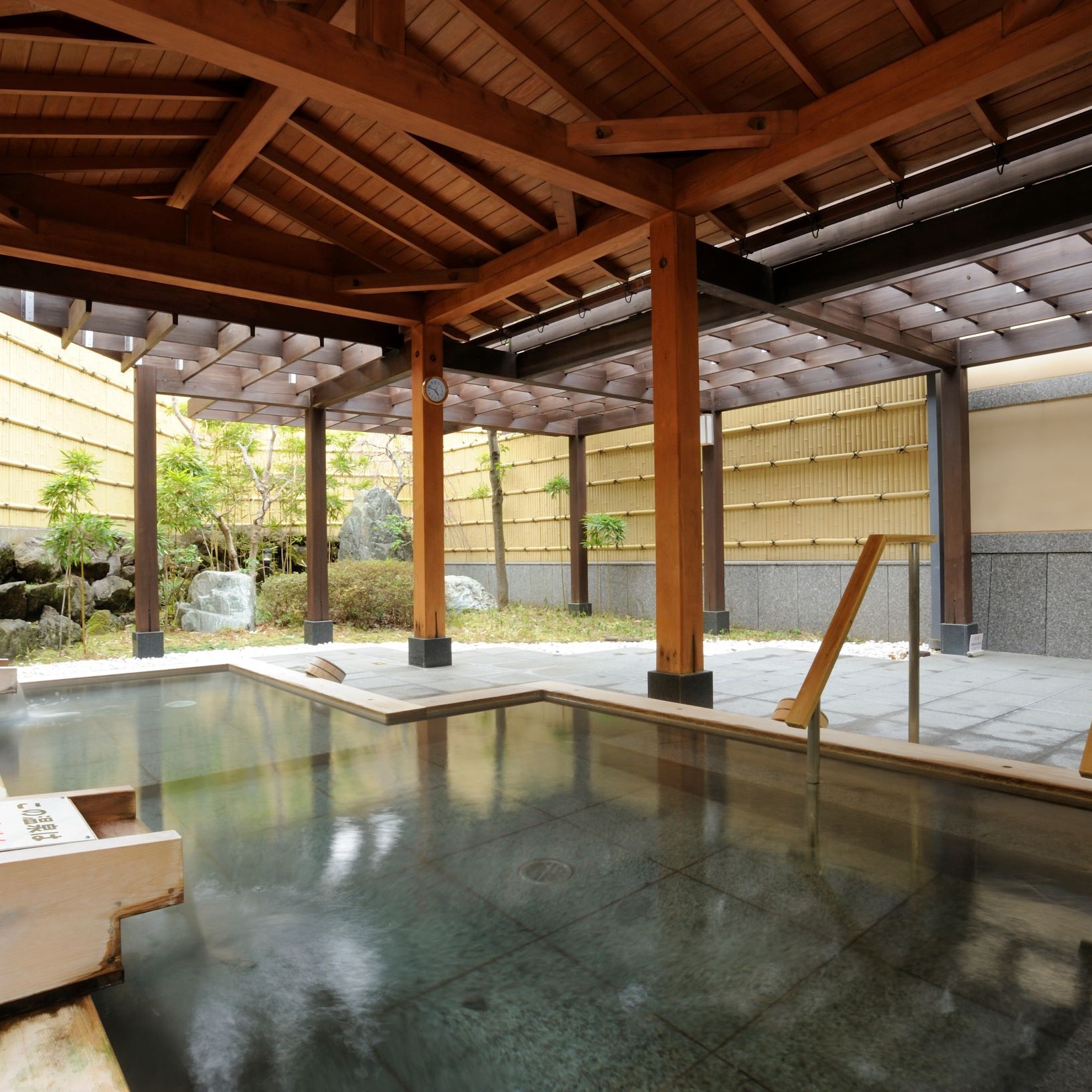 格子状の天井と太い木製の柱に支えられた屋根の下に、広い湯舟があり、その奥には竹垣と岩や木々が配された庭園が見える開放的な露天風呂の写真