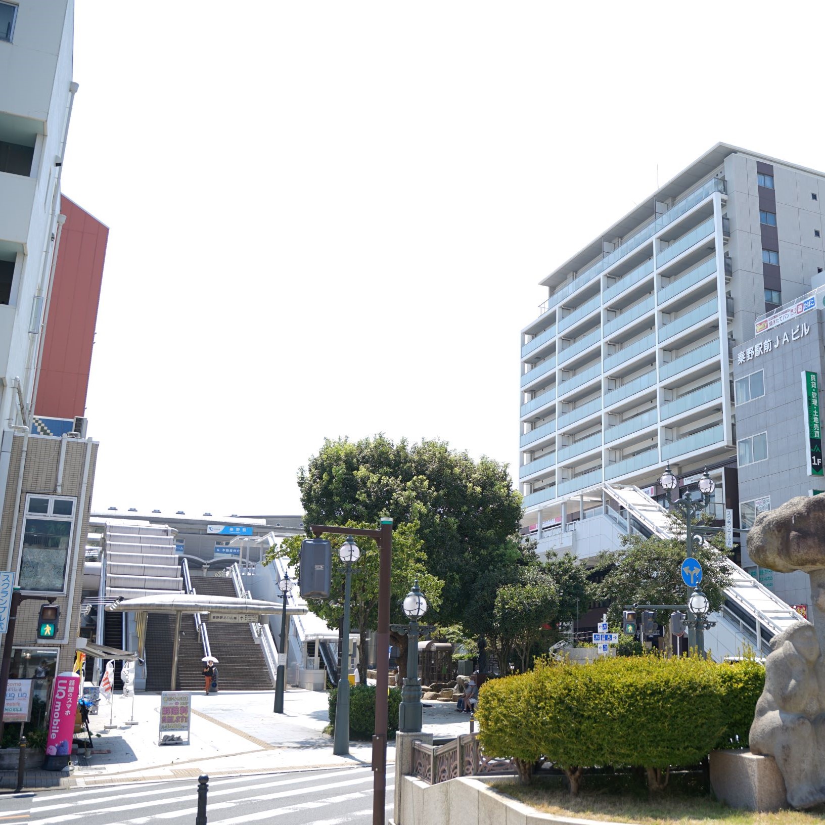 広場の奥に駅の階段とプラットフォーム、左には白い壁と赤い建物、右には「秦野駅前JAビル」と書かれたビルや大きなマンションが立つ、晴れた日の秦野駅北口ロータリー周辺の風景写真