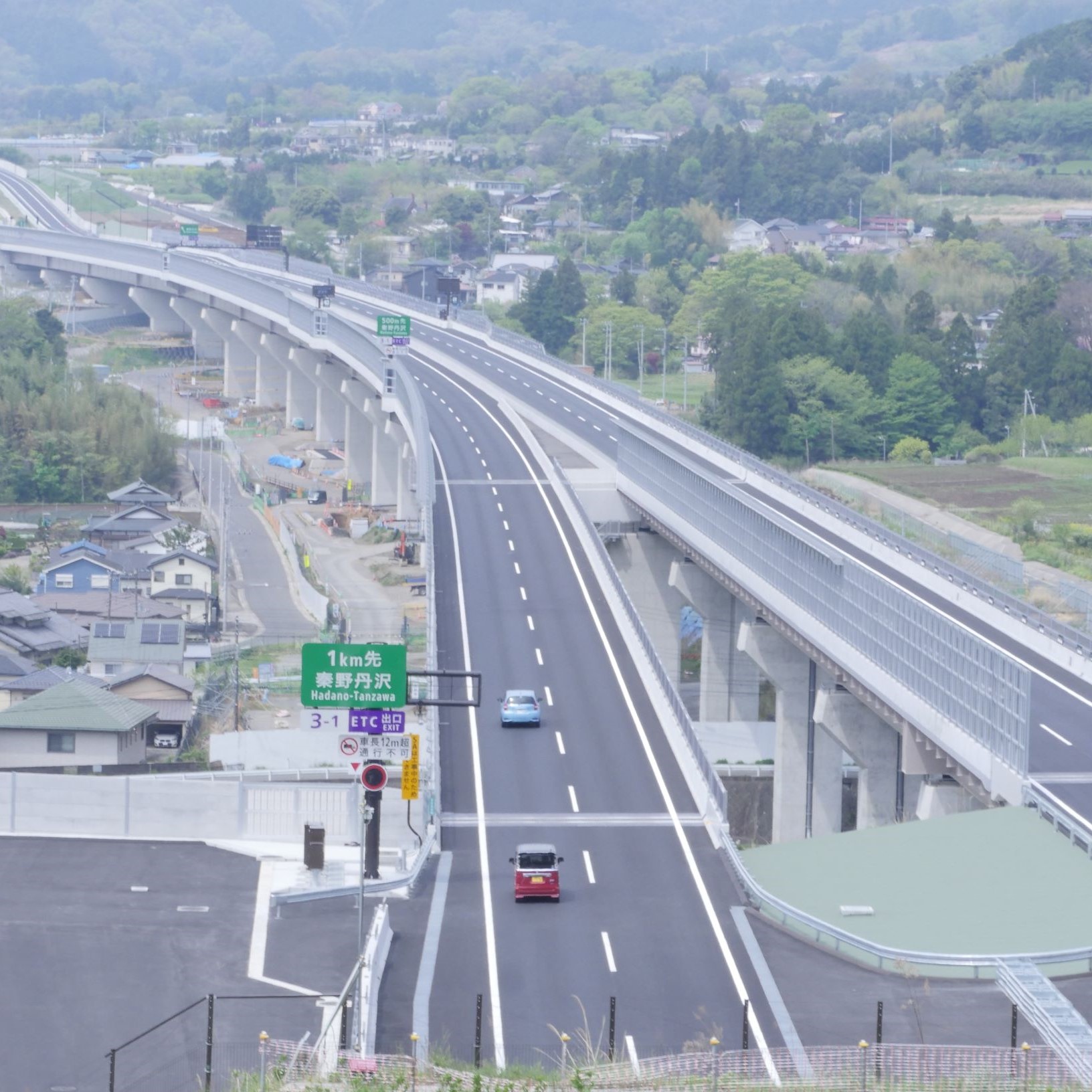 コンクリート製の高い高架橋が左右に伸び、その上を上下線が分かれて舗装され、左側手前には「 ETC専用 秦野丹沢」と書かれた緑の案内標識が見える高速道路の写真