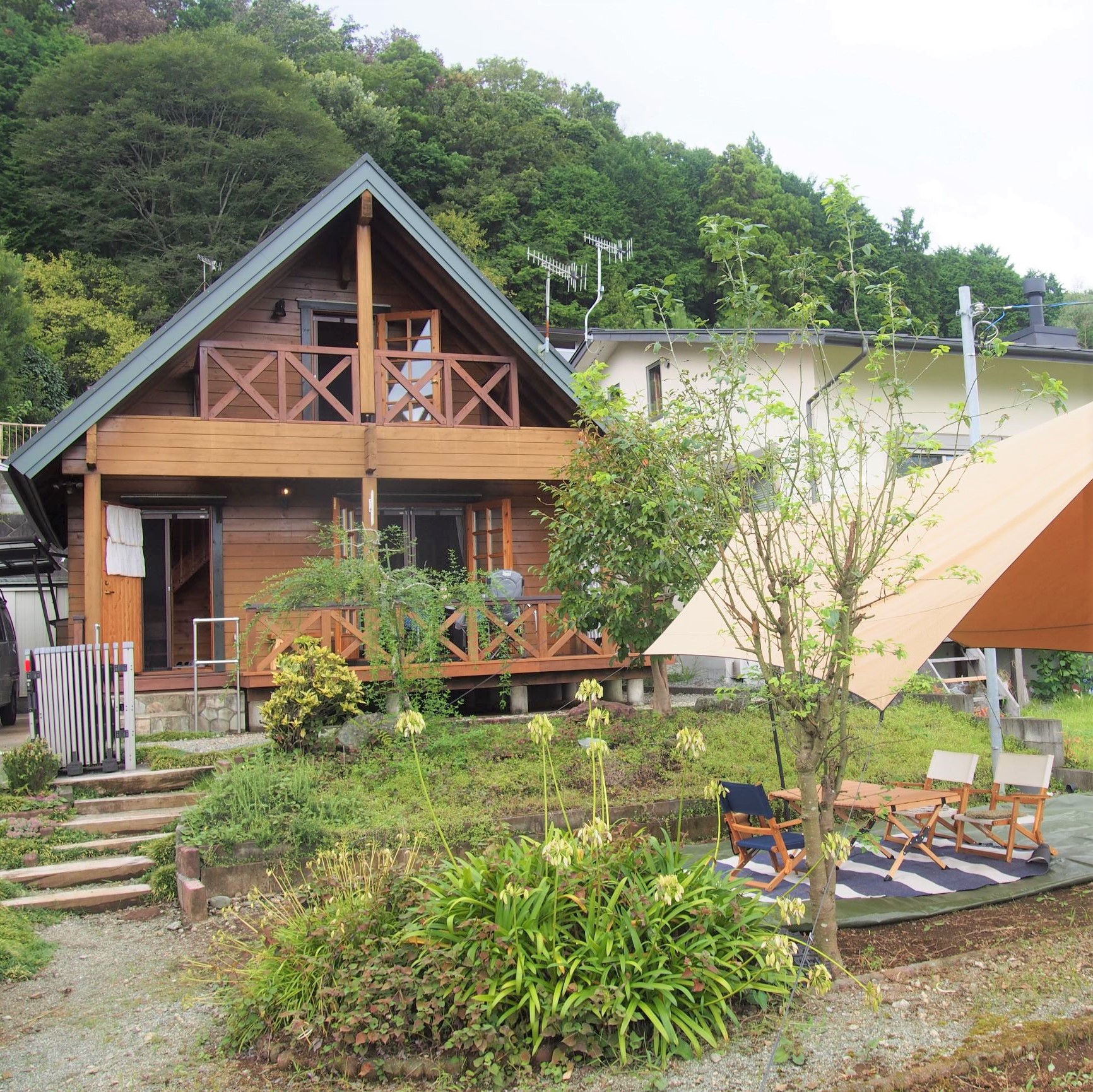 二階建ての木造ロッジの前に石段と広い庭があり、庭には植栽とタープの下に椅子とテーブルが置かれ、背景には豊かな緑の森が広がるコテージの外観写真