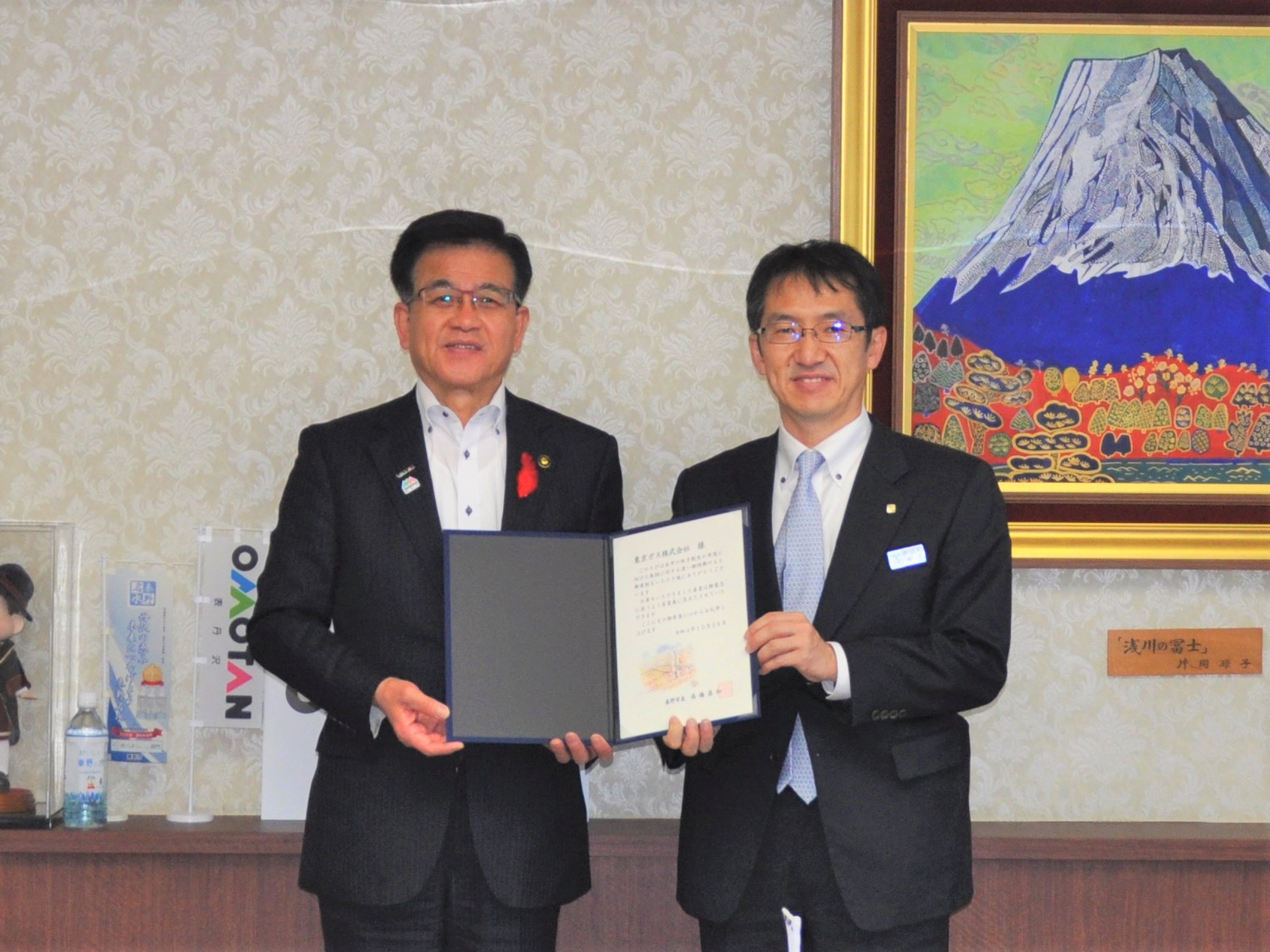 富士山の絵画を背景にした室内のカウンター前で、市長と東京ガス株式会社の関係者が、中央で証書のような書類を広げて持ち、カメラに向かって真剣な表情で立っている記念写真
