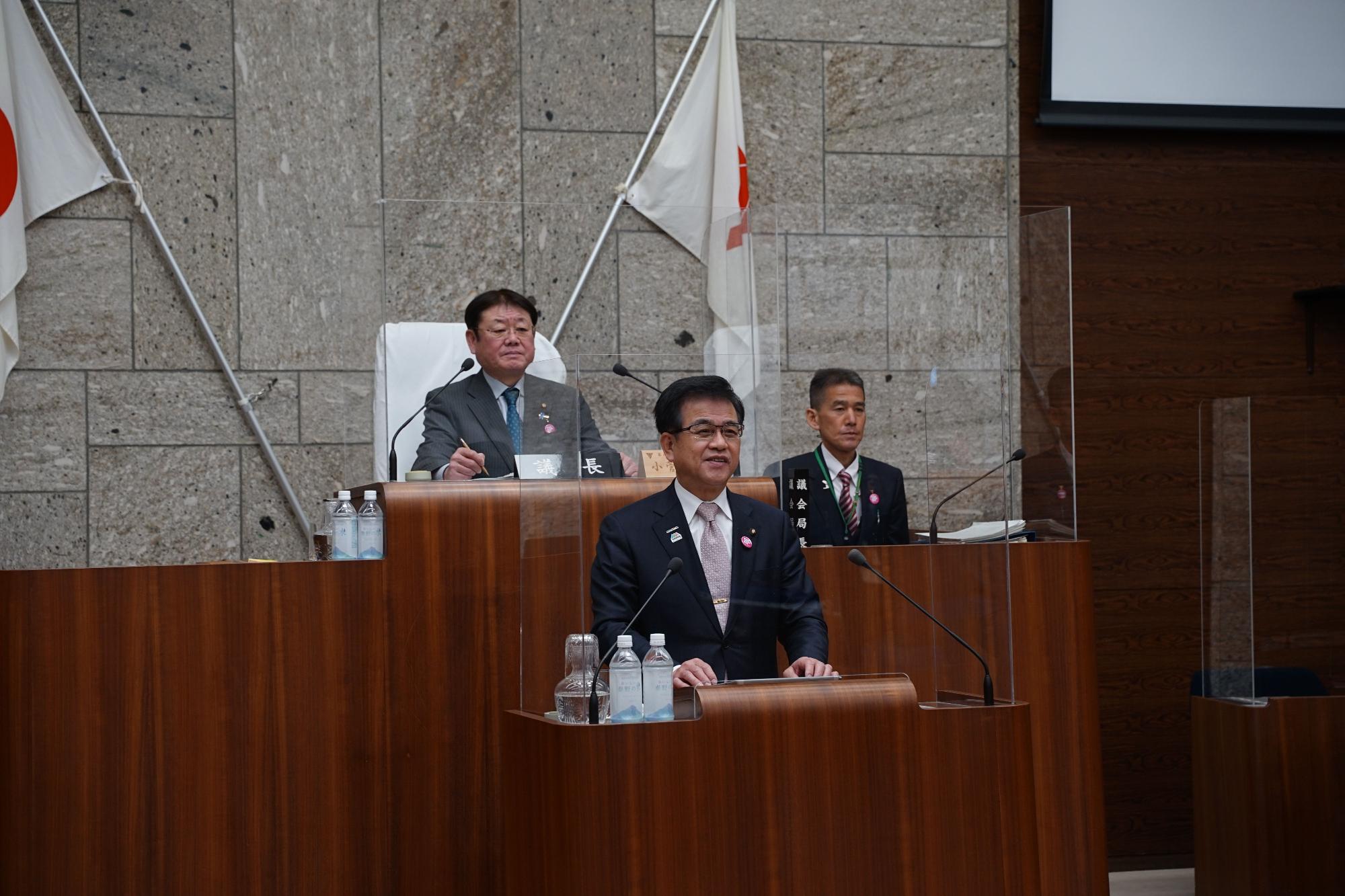 木製の演壇の前にダークスーツを着た市長が立ち、マイクに向かって穏やかな表情で話しており、その後ろの壇上には別の二人のスーツ姿の男性が着席し、周囲には日本の国旗と白い旗が立てられた会議の様子の写真