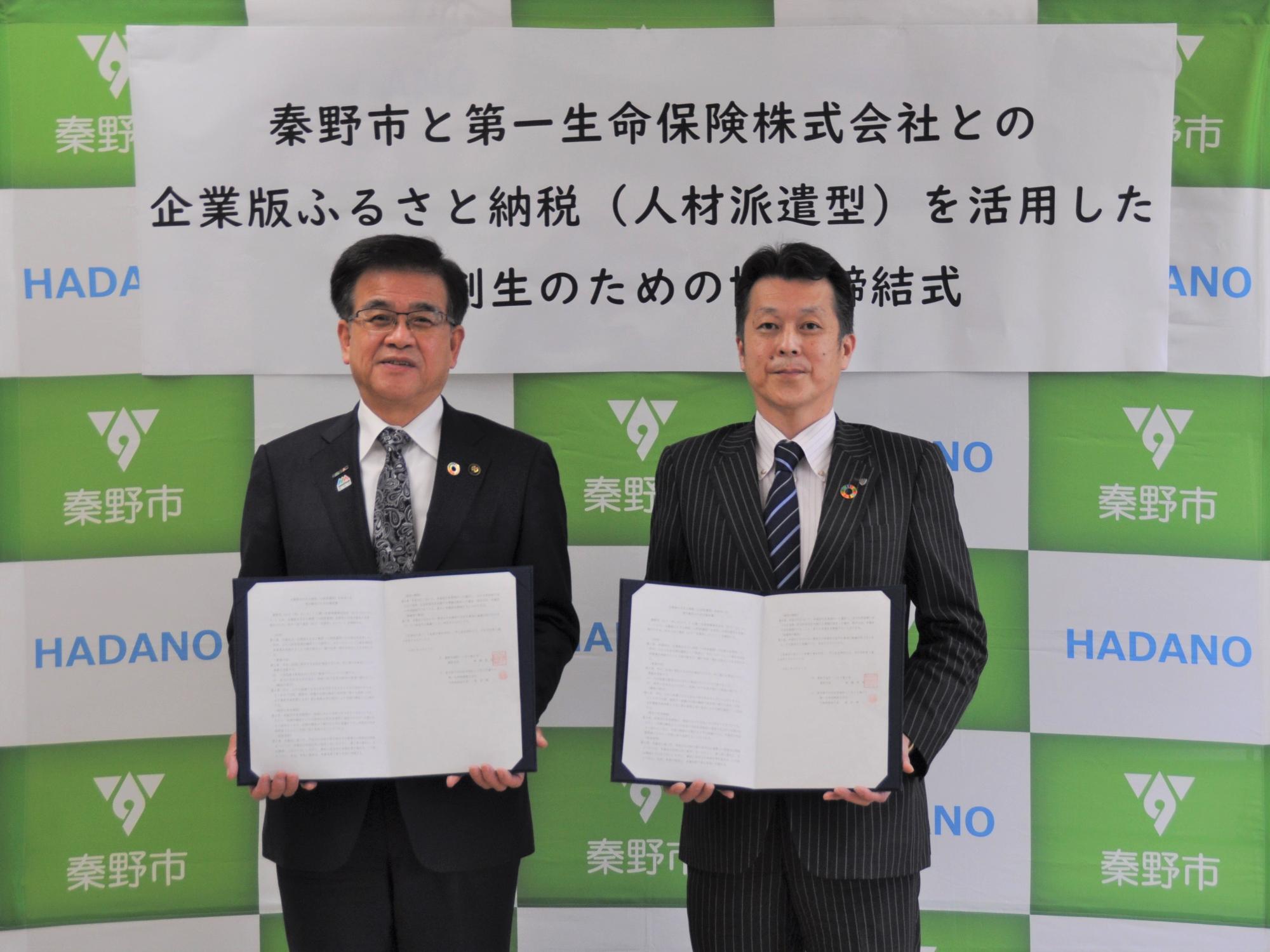 白と緑のロゴ入りの背景幕の前で、スーツ姿の市長と男性がそれぞれ開いた協定書を持って正面を向き、笑顔で立っている写真