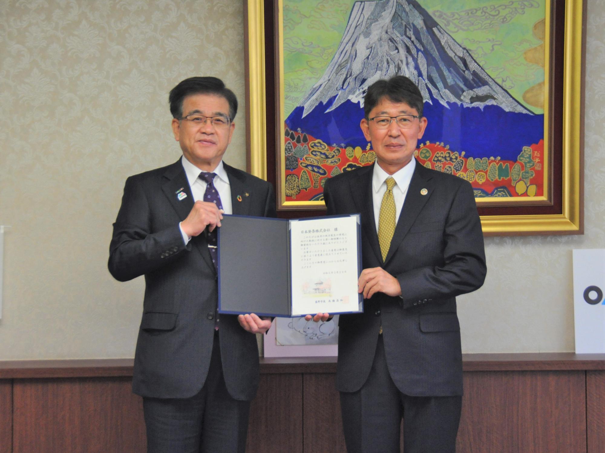 富士山の絵画を背景にした室内のカウンター前で、市長と株式会社トープラ様の関係者が、中央で証書のような書類を広げて持ち、カメラに向かって真剣な表情で立っている記念写真