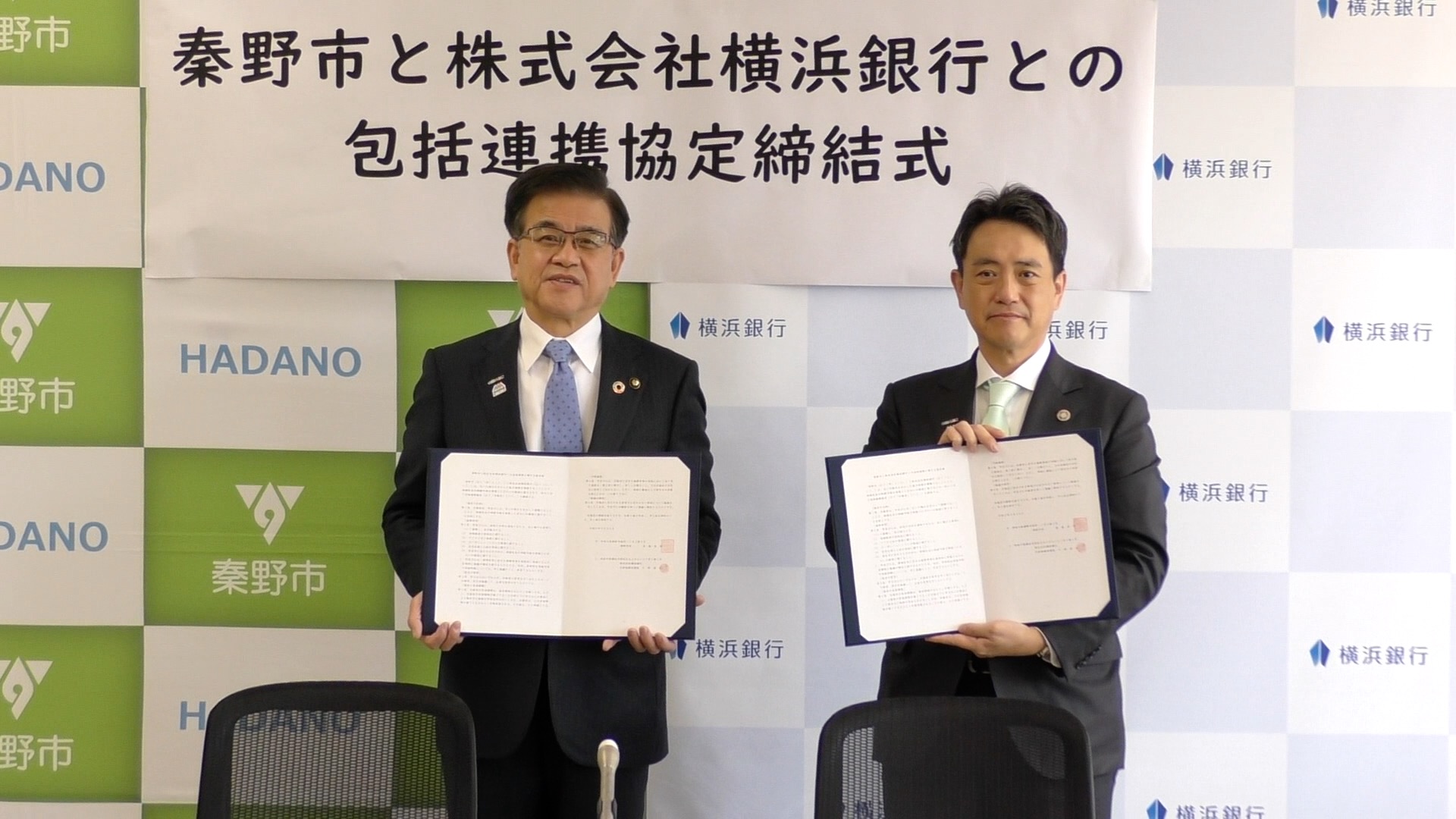 秦野市のバックボードの前で高橋市長と横浜銀行の男性が協定書を持ち記念撮影をしている写真